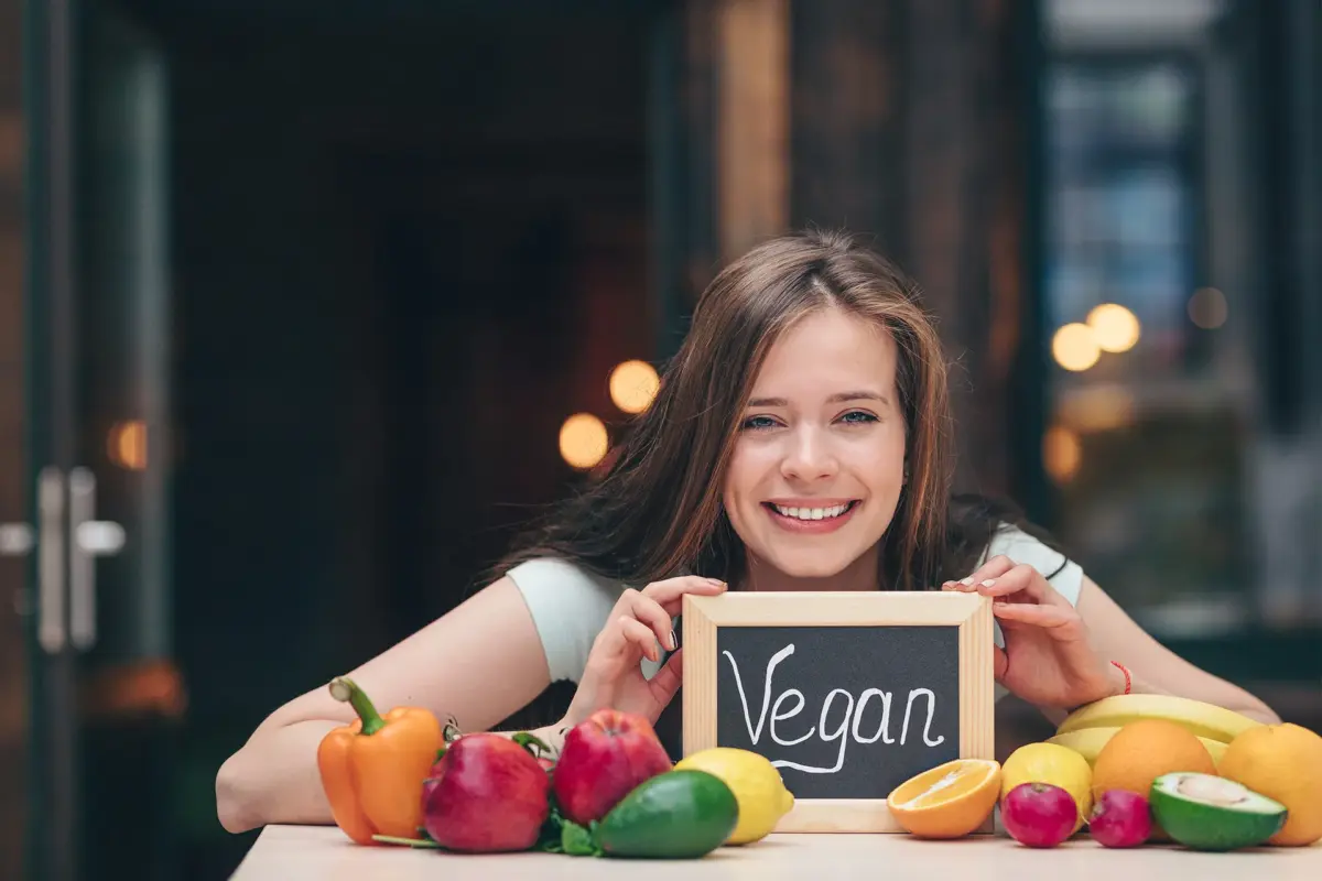 Junge Frau mit Tafel „Vegan“ vor buntem Obst und Gemüse – Symbolbild für vegane Ernährung und Nahrungsergänzung vegan