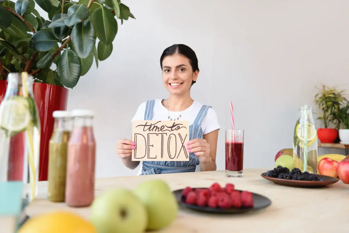 Frau mit Schild „Time to Detox“ sitzt am Tisch mit frischen Früchten und Smoothies