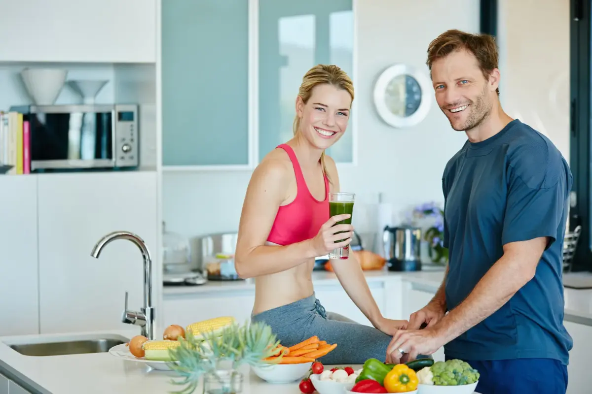 Sportliches Paar in der Küche mit Smoothie und frischem Gemüse als Symbol für Vitamine Abnehmen Unterstützung