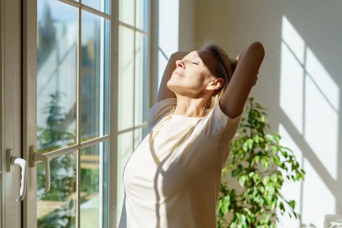 Frau steht am Fenster im Sonnenlicht und entspannt sich zur Unterstützung bei Frühjahrsmüdigkeit Ursachen