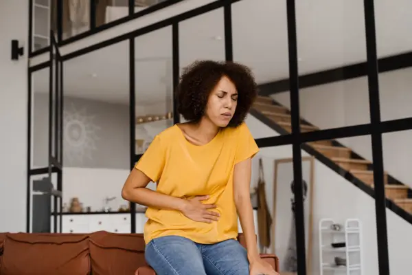 Junge Frau im gelben T-Shirt hält sich mit schmerzverzerrtem Gesicht den Bauch – Symbolbild für Blähungen Ursachen Ernährung