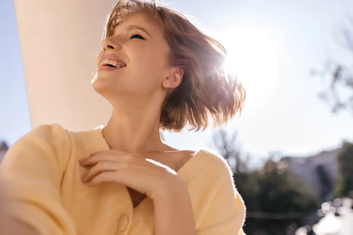 Junge Frau im gelben Cardigan lacht glücklich im warmen Sonnenlicht mit strahlender Haut – Symbolbild für Hautschutz von innen im Sommer