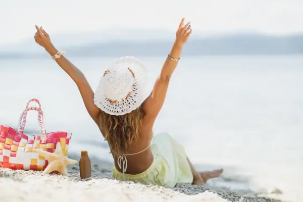 Junge Frau mit weißem Sonnenhut sitzt entspannt am Strand und streckt die Arme in die Höhe – Symbolbild für einen unbeschwerten Sommerurlaub mit ausgeglichener Verdauung