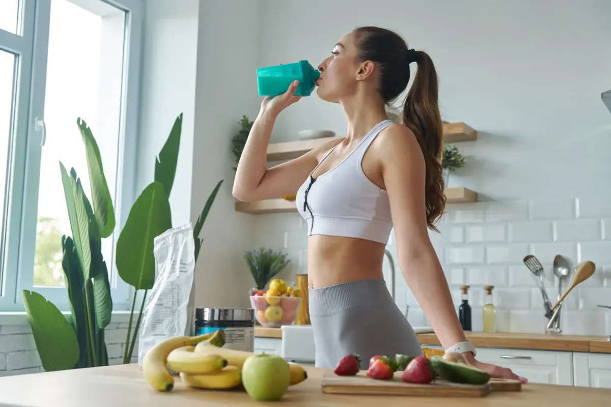 Sportliche Frau trinkt einen Proteinshake aus einem türkisen Shaker in einer hellen Küche – Symbolbild für die Wirkung von Proteinen im Körper