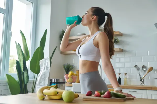 Sportliche Frau trinkt einen Proteinshake aus einem türkisen Shaker in einer hellen Küche – Symbolbild für die Wirkung von Proteinen im Körper