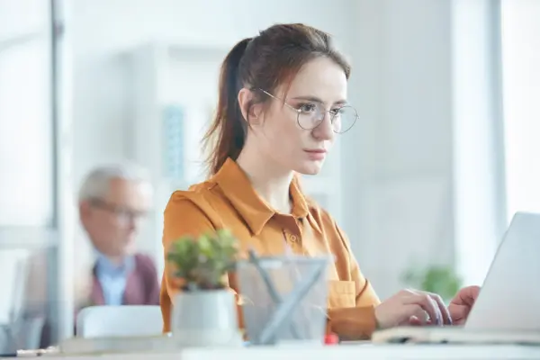 Junge Frau mit Brille arbeitet konzentriert am Laptop in einem hellen Büro – Konzentration steigern: natürlich mehr Fokus im Arbeitsalltag
